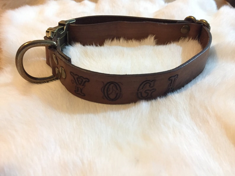 May include: A brown leather dog collar with a metal buckle and a large metal ring. The collar has the word "YOGI" embossed on it. The collar is designed to fit around a dog's neck.