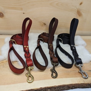 May include: Four leather dog leashes in red, brown, and black, each with a braided section and a metal clasp. The leashes are displayed on a wooden board with a white fur-like material.