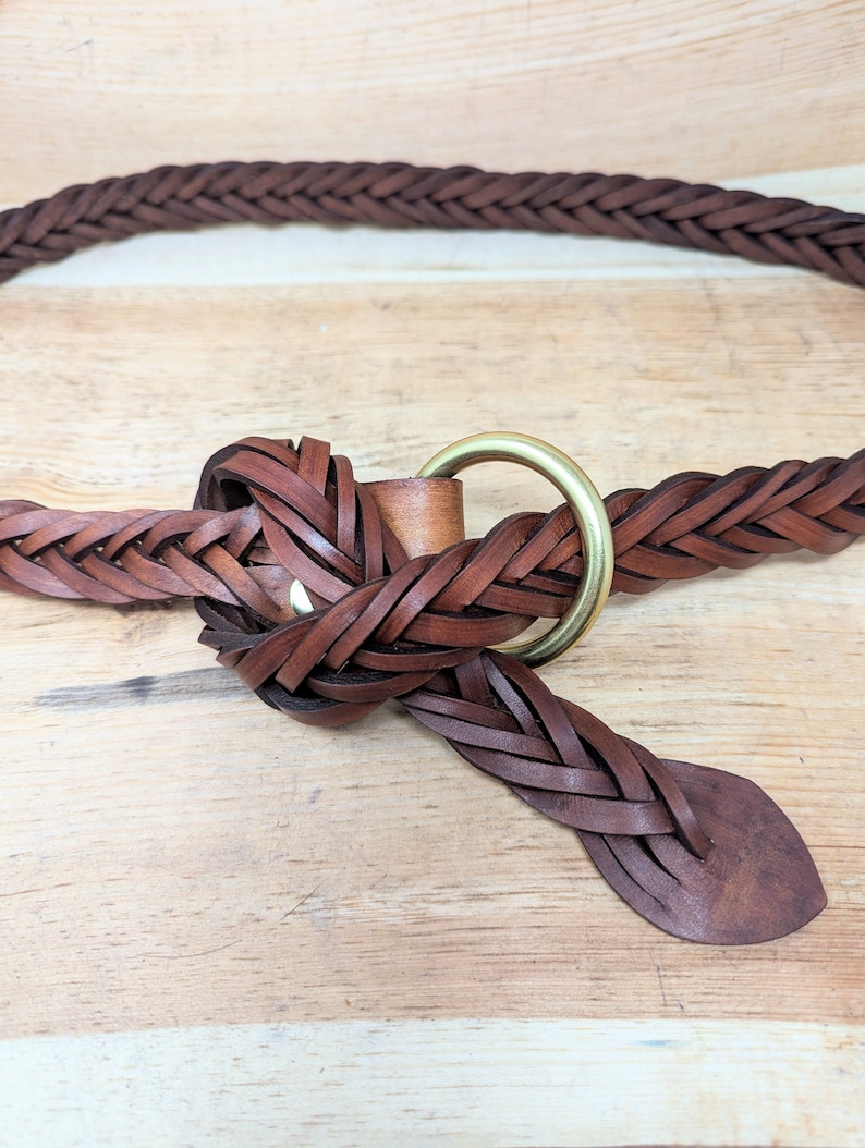 K&ouml;nnte beinhalten: Geflochtener brauner Lederg&uuml;rtel mit einer Messingringschlie&szlig;e. Der G&uuml;rtel besteht aus geflochtenen Lederstreifen und hat ein klassisches Design. Der G&uuml;rtel wird auf einer hellen Holzoberfl&auml;che gezeigt.