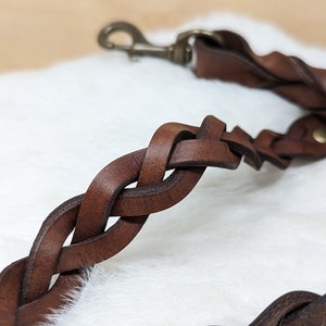 May include: Close-up of a braided brown leather dog leash with a brass-colored clasp. The leash is made of thick, interwoven leather strips, creating a durable and stylish design. The background is a soft, white, fluffy surface.