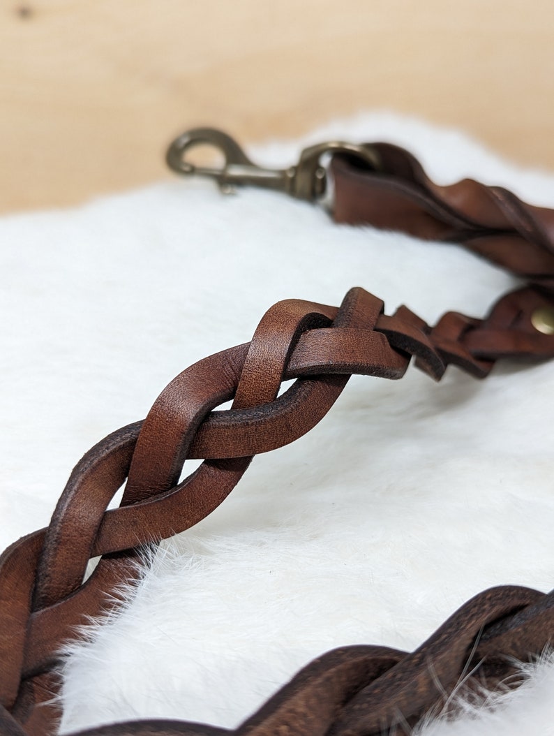 May include: Close-up of a braided brown leather dog leash with a brass-colored clasp. The leash is made of thick, interwoven leather strips, creating a durable and stylish design. The background is a soft, white, fluffy surface.