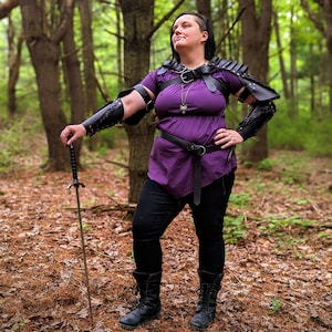 May include: A person wearing a purple tunic, black pants, and black boots, posing in a forest. The person is wearing black leather armor on the shoulders and arms, and holding a sword. A silver pendant hangs from a necklace.