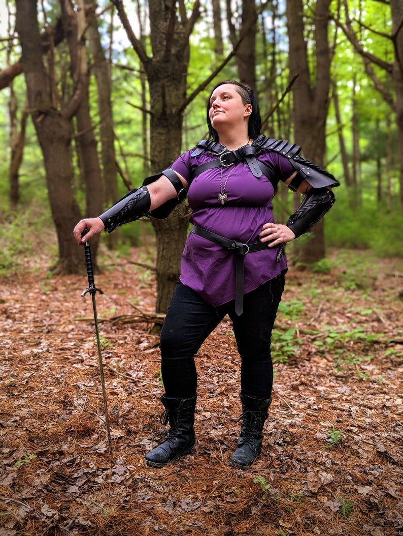 May include: A person wearing a purple tunic, black pants, and black boots, posing in a forest. The person is wearing black leather armor on the shoulders and arms, and holding a sword. A silver pendant hangs from a necklace.