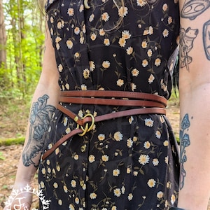 May include: A brown leather belt with a gold-colored ring buckle is wrapped around a black dress with a white and yellow floral pattern. The belt is worn over the dress, cinching the waist. The dress has a button-down front.