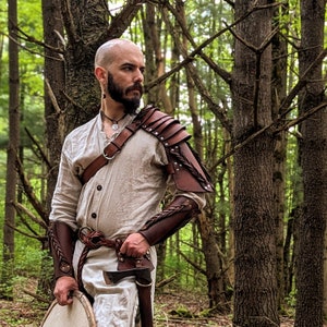 May include: A person wearing a light-colored tunic and leather armor, including a shoulder pauldron and arm bracers. The armor is brown with decorative braiding and metal studs. The person is holding a small axe and a round shield.