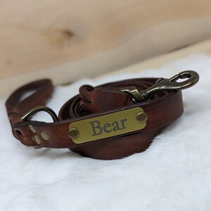 May include: A rolled brown leather dog leash with a brass clasp and a brass nameplate that reads "Bear". The leash has a loop handle and is lying on a white, fluffy surface.