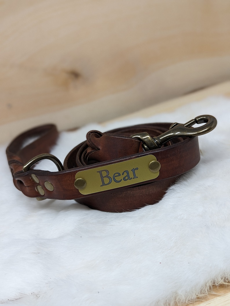 May include: A rolled brown leather dog leash with a brass clasp and a brass nameplate that reads "Bear". The leash has a loop handle and is lying on a white, fluffy surface.