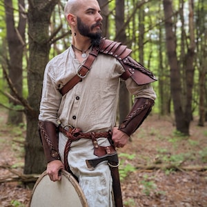 K&ouml;nnte beinhalten: Eine Person tr&auml;gt eine beigefarbene Tunika und Lederpanzerung, einschlie&szlig;lich Schulterpolstern und Armsch&uuml;tzern. Das Outfit ist mit einem geflochtenen G&uuml;rtel, einer kleinen Axt und einem runden Schild ausgestattet. Der Hintergrund ist ein Wald.