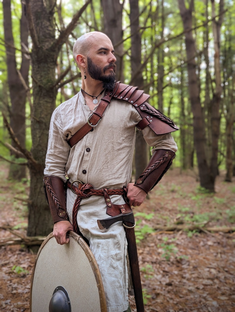 K&ouml;nnte beinhalten: Eine Person tr&auml;gt eine beigefarbene Tunika und Lederpanzerung, einschlie&szlig;lich Schulterpolstern und Armsch&uuml;tzern. Das Outfit ist mit einem geflochtenen G&uuml;rtel, einer kleinen Axt und einem runden Schild ausgestattet. Der Hintergrund ist ein Wald.