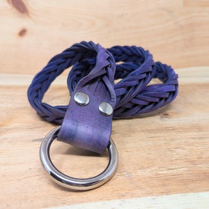 K&ouml;nnte beinhalten: Ein geflochtener G&uuml;rtel in Indigo- und Lilat&ouml;nen mit einem gro&szlig;en, silberfarbenen Metallring. Der G&uuml;rtel besteht aus gewebten Materialstreifen und ist mit zwei silbernen Nieten befestigt. Der G&uuml;rtel wird auf einer hellen Holzoberfl&auml;che pr&auml;sentiert.