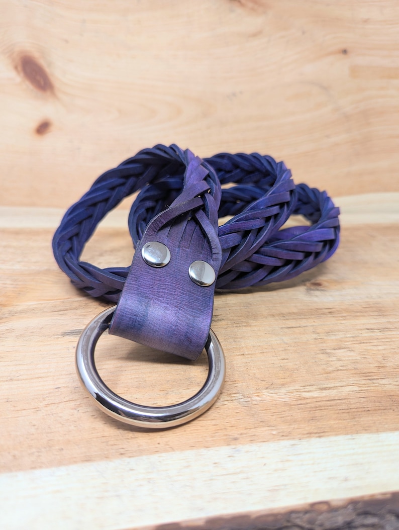 K&ouml;nnte beinhalten: Ein geflochtener G&uuml;rtel in Indigo- und Lilat&ouml;nen mit einem gro&szlig;en, silberfarbenen Metallring. Der G&uuml;rtel besteht aus gewebten Materialstreifen und ist mit zwei silbernen Nieten befestigt. Der G&uuml;rtel wird auf einer hellen Holzoberfl&auml;che pr&auml;sentiert.