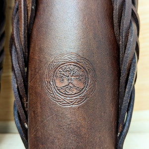 May include: Close-up of a brown leather item with a braided strap. The item features a circular embossed design of a tree with intertwined roots and branches. A decorative border is embossed at the bottom.