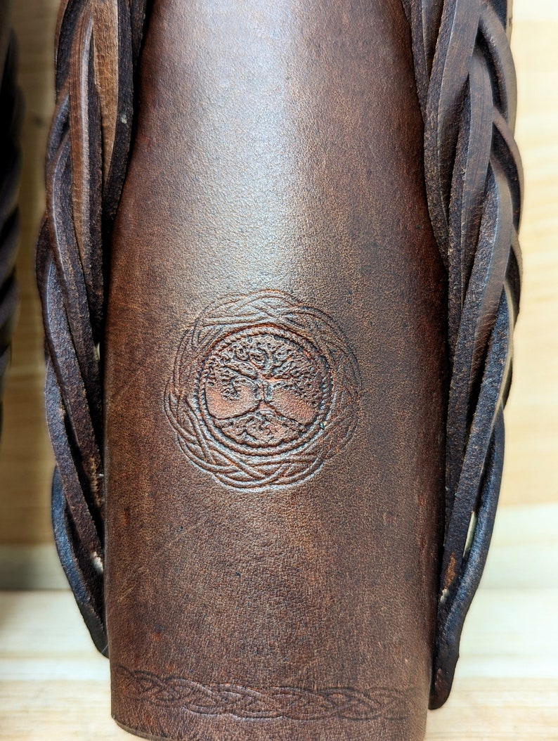 May include: Close-up of a brown leather item with a braided strap. The item features a circular embossed design of a tree with intertwined roots and branches. A decorative border is embossed at the bottom.