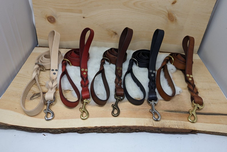May include: A collection of leather dog leashes in various colors, including tan, red, brown, black, and dark brown. Each leash features a braided handle and a metal clasp. The leashes are displayed on a wooden surface.