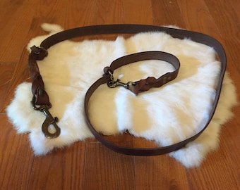 May include: A brown leather dog leash and matching collar with brass-colored hardware. The leash features a braided section near the clasp. The items are displayed on a white, fluffy surface.