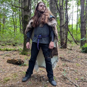 K&ouml;nnte beinhalten: Eine Person in einem mittelalterlichen Outfit steht in einem Wald. Das Outfit umfasst einen pelzbesetzten Umhang, eine graue Tunika mit einem lila Seilg&uuml;rtel, schwarze Hosen und schwarze Stiefel. Der Umhang hat einen blauen Akzent. Die Person hat lange braune Haare.