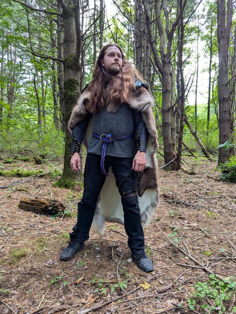K&ouml;nnte beinhalten: Eine Person in einem mittelalterlichen Outfit steht in einem Wald. Das Outfit umfasst einen pelzbesetzten Umhang, eine graue Tunika mit einem lila Seilg&uuml;rtel, schwarze Hosen und schwarze Stiefel. Der Umhang hat einen blauen Akzent. Die Person hat lange braune Haare.