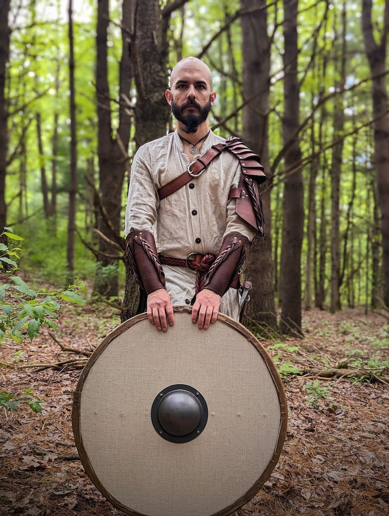 May include: A person wearing a cream-colored tunic, brown leather armor, and holding a round shield. The shield has a light-colored fabric face with a dark metal boss in the center and a wooden frame. The background is a forest.
