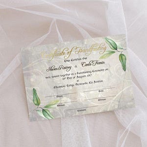 May include: A certificate with gold script that reads "Certificate of Handfasting". The certificate has a floral design and green leaf accents. The names Steven Rainey and Carla Ferrin are printed on the certificate.