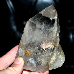 Puede incluir: Un cristal de cuarzo transparente con una punta y una superficie rugosa y texturizada. El cristal tiene inclusiones de minerales negros y dorados. El cuarzo se sostiene en una mano sobre un fondo oscuro.