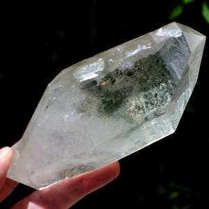 Puede incluir: Un cristal de cuarzo transparente con una punta. El cristal tiene una apariencia translúcida con algunas inclusiones internas. El cristal se sostiene en una mano sobre un fondo oscuro. El cristal es un mineral natural.