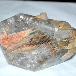 May include: A clear quartz crystal with internal inclusions of reddish-orange, brown, and gold hues. The crystal has a multifaceted shape and a rough, natural texture. A smaller quartz formation is attached to the top of the main crystal.