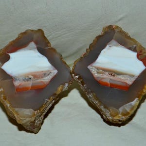 Puede incluir: Dos mitades de una geoda, que muestran una formación de piedra natural. El interior presenta un patrón blanco, naranja y gris, enmarcado por un borde exterior marrón y beige. La superficie rugosa y texturizada se suma a la estética natural.