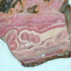 Puede incluir: Una losa pulida de piedra de rodocrosita, que muestra capas de tonos rosa, blanco y marrón rojizo. La piedra presenta patrones circulares concéntricos y un borde natural rugoso, creando una pieza decorativa única.