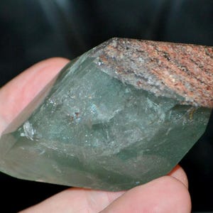 Puede incluir: Cristal translúcido verde con una parte superior rugosa de color marrón rojizo. El cristal tiene una forma facetada, con estructuras internas visibles y una calidad ligeramente iridiscente. La apariencia general sugiere un espécimen mineral natural.