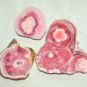 Puede incluir: Tres piezas de piedra preciosa de rodocrosita. Las piedras muestran bandas concéntricas de color rosa, rojo y blanco, con un aspecto en capas. La pieza más grande tiene una forma redondeada e irregular, mientras que las otras están cortadas.