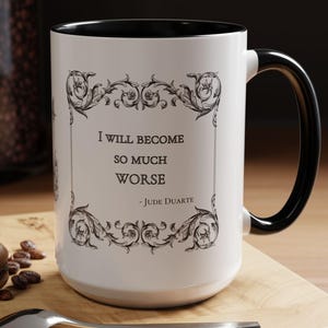 May include: White ceramic mug with a black interior and handle. The mug features the quote "I WILL BECOME SO MUCH WORSE - Jude Duarte" in black text within a decorative frame. A spoon and a macaron are on a wooden surface.