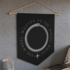 Peut inclure: Une tenture murale noire avec le texte "THE WHEEL WEAVES AS THE WHEEL WILLS" en blanc. Le design présente un ouroboros, un serpent se mordant la queue, et une étoile. La bannière est suspendue à une tringle en bois.