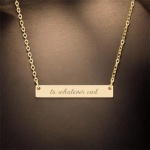 May include: A gold bar necklace with the inscription "to whatever end". The necklace has a fine chain and a rectangular pendant. The jewellery is displayed against a dark background. The pendant is engraved with a cursive font.