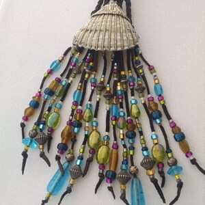 May include: A long necklace with a silver seashell pendant and a fringe of colorful beads. The beads are in shades of blue, green, brown, and gold. The necklace is made of brown leather cord.
