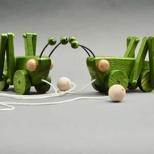 May include: Two green wooden toy grasshoppers connected by a white string. Each grasshopper has four wheels and a pair of antennae. The grasshoppers are facing each other.