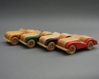 May include: Five wooden toy cars lined up in a row. The cars are painted in different colors, including yellow, green, black, and red. The cars are made of natural wood and have a simple, classic design.