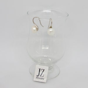 May include: A pair of silver dangle earrings with white pearl accents. The earrings are displayed on a clear glass pedestal with a "JZ Jewelry" label.