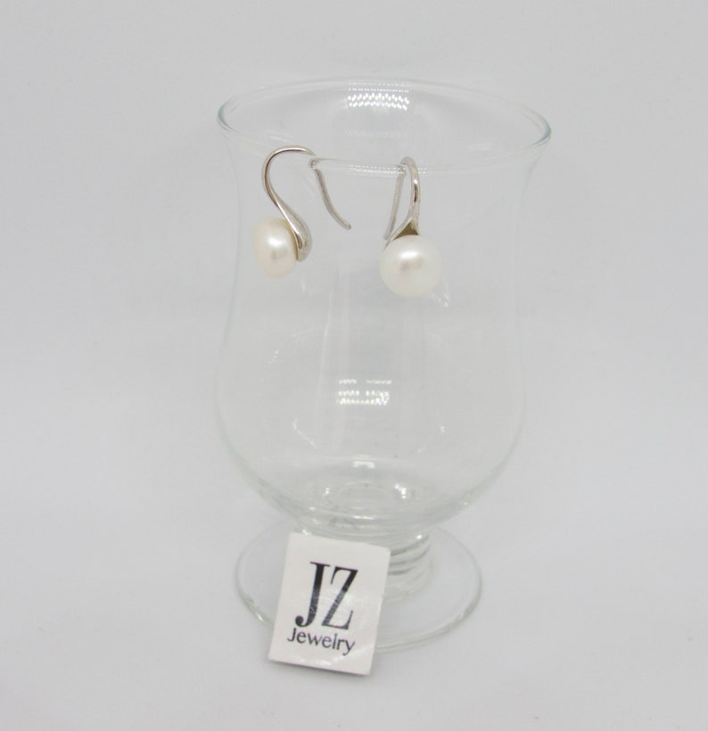 May include: A pair of silver dangle earrings with white pearl accents. The earrings are displayed on a clear glass pedestal with a "JZ Jewelry" label.
