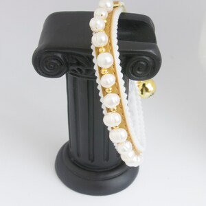 May include: A white and gold cat collar with a row of white pearls and a gold bell. The collar is displayed on a black pedestal.