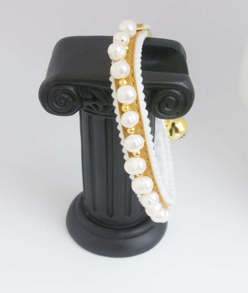 May include: A white and gold cat collar with a row of white pearls and a gold bell. The collar is displayed on a black pedestal.
