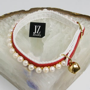 May include: A red and white leather cat collar with a row of white pearls and a gold bell. The collar has a white leather lining and a black tag that reads "JZ Jewelry".