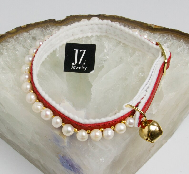 May include: A red and white leather cat collar with a row of white pearls and a gold bell. The collar has a white leather lining and a black tag that reads "JZ Jewelry".