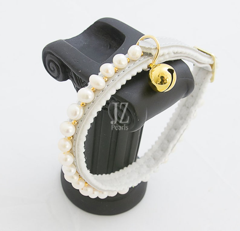 May include: White leather cat collar with a row of white pearls and a gold bell. The collar is on a black stand. "JZ Pearls" is printed on the stand.