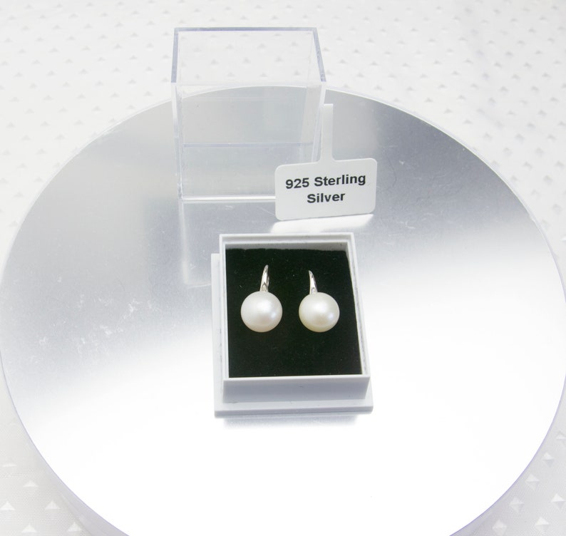 May include: A pair of white pearl earrings with sterling silver hooks in a white box with a black interior. The box has a label that reads "925 Sterling Silver".