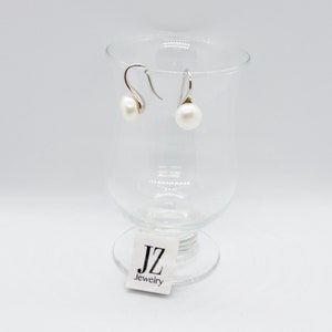 May include: A pair of silver dangle earrings with white pearl drops. The earrings are displayed on a clear glass pedestal with a white label that reads "JZ Jewelry".
