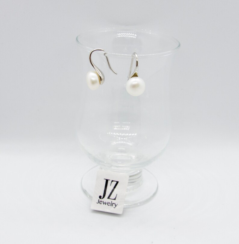 May include: A pair of silver dangle earrings with white pearl drops. The earrings are displayed on a clear glass pedestal with a white label that reads "JZ Jewelry".