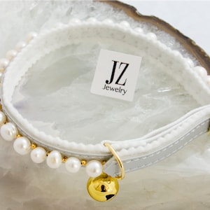 May include: White cat collar with a row of white pearls and a gold bell. The collar is made of white leather with a white fleece lining. The collar has a gold buckle and a gold ring for attaching a leash. The collar is labeled "JZ Jewelry".