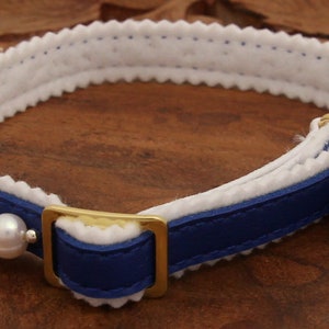 May include: A blue leather cat collar with a white felt lining and a row of white pearls. The collar has a gold buckle and a gold bell.