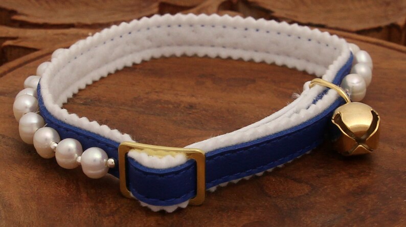 May include: A blue leather cat collar with a white felt lining and a row of white pearls. The collar has a gold buckle and a gold bell.