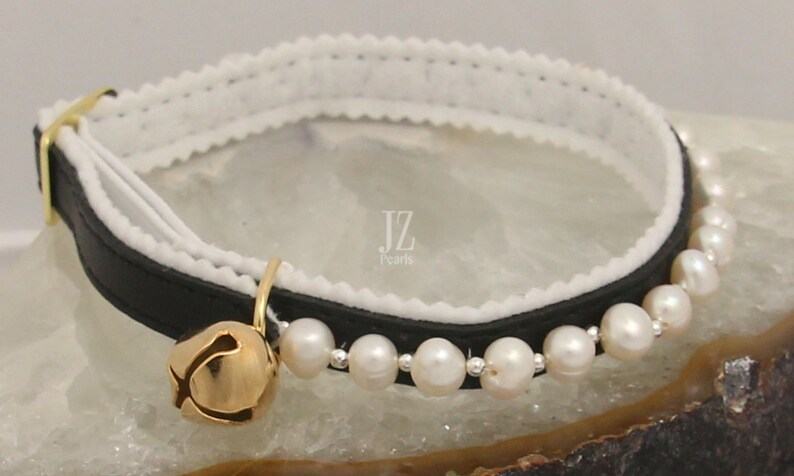 May include: Black leather cat collar with a white velvet lining and a row of white pearls. The collar has a gold bell attached.